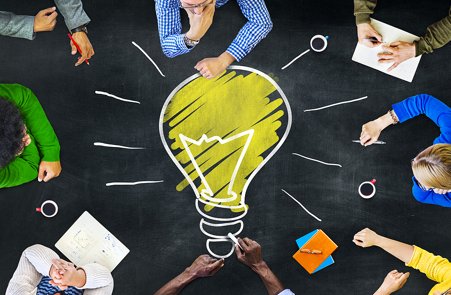 Image of a roughly drawn incandescent light-bulb on a chalk-board like table. Sitting around the lightbulb are eight people in various poses of study, thinking and note-taking.